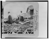 Two women stand amidst debris and rubble from destroyed buildings, possibly searching for remnants of their home. The scene depicts widespread destruction with wooden planks scattered around, and a partially standing house in the background.