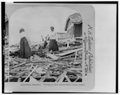 Two women stand amidst debris and rubble from destroyed buildings, possibly searching for remnants of their home. The scene depicts widespread destruction with wooden planks scattered around, and a partially standing house in the background.
