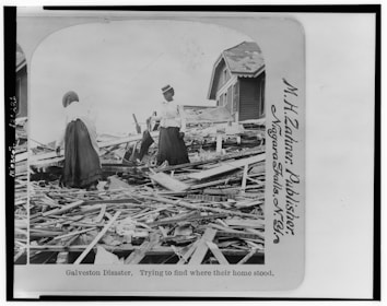Two women stand amidst debris and rubble from destroyed buildings, possibly searching for remnants of their home. The scene depicts widespread destruction with wooden planks scattered around, and a partially standing house in the background.