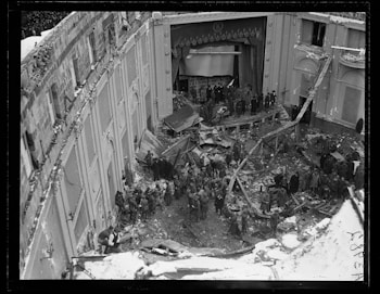 A theater interior is heavily damaged, with debris scattered across the stage and seating area. Several people, possibly emergency responders or military personnel, are present amidst the wreckage. The architectural details, like ornate decorations and large curtains, are visible on the stage. The entire scene conveys a sense of destruction and organized response effort.
