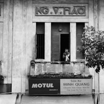 An old building with a person sitting on the windowsill, looking outward contemplatively. The architecture is weathered with large shutters and a faded sign with Vietnamese text beneath the window. A tree partially obscures the view on the right side.