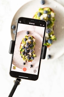 A smartphone is mounted on a tripod, capturing a close-up shot of a slice of toast topped with avocado, crumbled cheese, and blueberries. The toast and toppings have vibrant colors, and the image on the phone screen mirrors the food seen in the background.