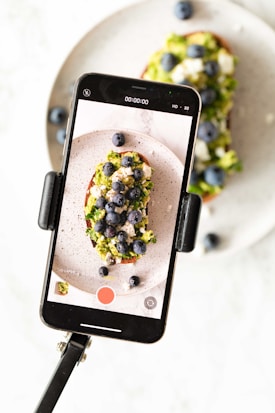 A smartphone is mounted on a tripod, capturing a close-up shot of a slice of toast topped with avocado, crumbled cheese, and blueberries. The toast and toppings have vibrant colors, and the image on the phone screen mirrors the food seen in the background.
