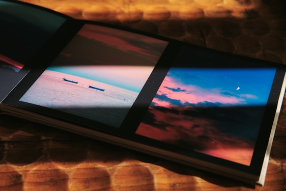 A photo album is open to two pages displaying picturesque skies. The left page shows a serene ocean scene with two boats floating on calm water, under a sky colored with hues of pink and blue. The right page captures a vivid sky during sunset with dramatic clouds and a crescent moon peeking through.