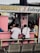 A small eatery with a pink exterior where several people are seated at a counter. The eatery, named 'Mommy J Eatery', is located at a stall and offers special dishes like lomi and silog. Inside, there are various bottles and items on shelves. The seating area includes benches and metal chairs.