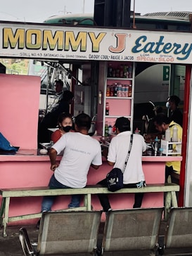 A small eatery with a pink exterior where several people are seated at a counter. The eatery, named 'Mommy J Eatery', is located at a stall and offers special dishes like lomi and silog. Inside, there are various bottles and items on shelves. The seating area includes benches and metal chairs.