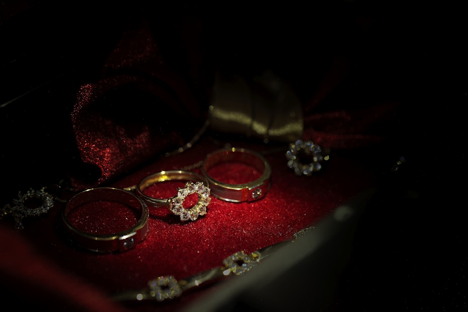 A collection of gold rings with various designs, embellished with small diamonds, is laid against a rich, dark red velvet fabric. The setting is dimly lit, emphasizing the sparkle of the jewels and the texture of the fabric.