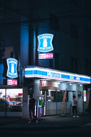 A Lawson convenience store is illuminated by bright neon signs at night. The store has blue and white signage with the Lawson logo prominently displayed. Two people are visible outside, one walking and another standing near the entrance. The store appears to be open, with lights shining from inside and various advertisements on display.