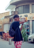 A person holding a camera in an urban setting, wearing a black t-shirt, red floral shorts, and a backpack. The backdrop includes a building with signage and outdoor restaurant seating.