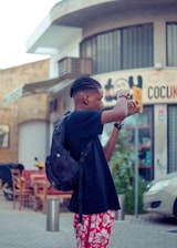 A person holding a camera in an urban setting, wearing a black t-shirt, red floral shorts, and a backpack. The backdrop includes a building with signage and outdoor restaurant seating.