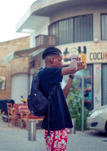 A person holding a camera in an urban setting, wearing a black t-shirt, red floral shorts, and a backpack. The backdrop includes a building with signage and outdoor restaurant seating.