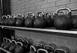 A row of black kettlebells with silver handles neatly arranged on a two-tier metal shelf against a brick wall. Each kettlebell is labeled with its weight in kilograms.