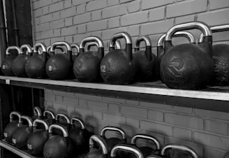 A row of black kettlebells with silver handles neatly arranged on a two-tier metal shelf against a brick wall. Each kettlebell is labeled with its weight in kilograms.