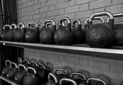 A row of black kettlebells with silver handles neatly arranged on a two-tier metal shelf against a brick wall. Each kettlebell is labeled with its weight in kilograms.