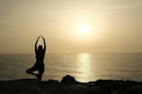 A person is practicing yoga, balanced on one leg with arms raised, silhouetted against a serene sunset over the ocean. The calm sea reflects the warm, golden hues of the setting sun.