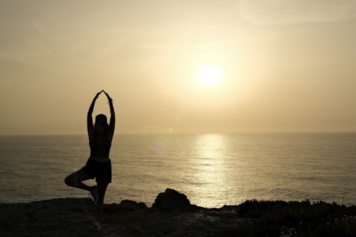 A person is practicing yoga, balanced on one leg with arms raised, silhouetted against a serene sunset over the ocean. The calm sea reflects the warm, golden hues of the setting sun.
