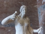 A marble statue of a woman with long, wavy hair holds objects in both hands, set against a textured stone wall background.
