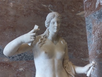 A marble statue of a woman with long, wavy hair holds objects in both hands, set against a textured stone wall background.