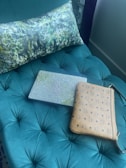 A green tufted cushion is adorned with a patterned pillow and accompanied by a textured laptop sleeve and a brown patterned clutch, all arranged on the cushion.
