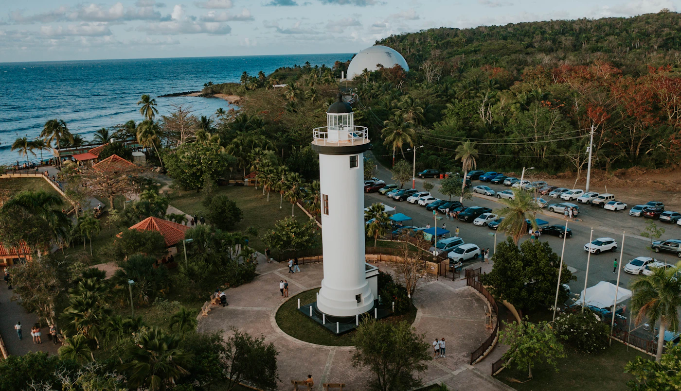 The Rincon lighthouse on the west coast of Puerto Rico on a rocky headland above the ocean with the Pacific sunset visible behind it and surfers and the ocean below