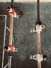 Several foosball players are positioned on metal rods on a foosball table. The players are wearing red and white uniforms, and the table surface has scuff marks indicating usage. The perspective is from above, capturing a section of the foosball field with visible white lines on a dark surface.
