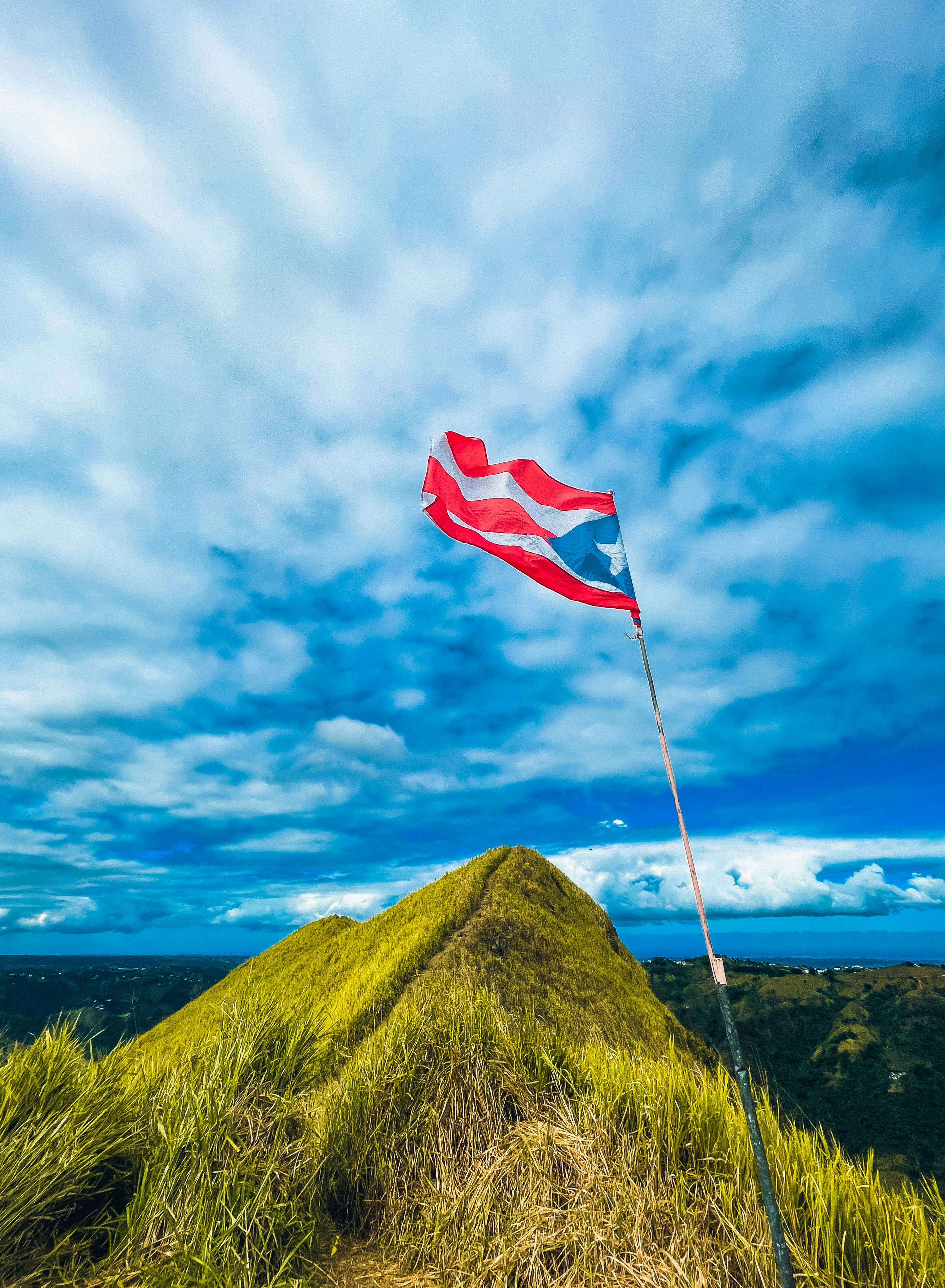 The Puerto Rican flag, its red and white stripes radiant next to the blue triangle that contains its star, flies above a grassy hill, looking over the greenery of the island against the blue sky.