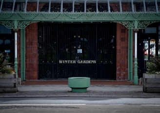 An ornate metal entrance with intricate detailing painted in pastel green stands before a brick wall. The words 'Winter Gardens' are displayed on the gates. Large planters with greenery are positioned on either side of the entrance. A circular green structure is situated in the foreground, and windows with reflections are visible on both sides.