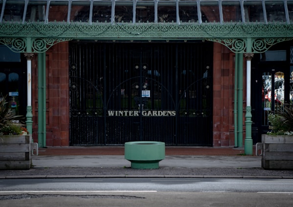 An ornate metal entrance with intricate detailing painted in pastel green stands before a brick wall. The words 'Winter Gardens' are displayed on the gates. Large planters with greenery are positioned on either side of the entrance. A circular green structure is situated in the foreground, and windows with reflections are visible on both sides.