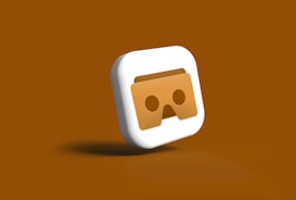 A three-dimensional icon featuring a stylized pair of VR goggles in the center. The goggles appear in a brownish shade on a square-shaped background with smooth, rounded edges. The icon is set against a monochromatic brown backdrop, enhancing the visual focus on the goggles.