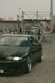 A sleek, dark-colored sports car with shiny rims is parked on a paved surface. Behind the car, there are metal barricades and scaffolding structures, with several people in the background. The setting appears to be an outdoor event or construction site, with various industrial elements visible.