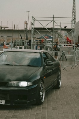 A sleek, dark-colored sports car with shiny rims is parked on a paved surface. Behind the car, there are metal barricades and scaffolding structures, with several people in the background. The setting appears to be an outdoor event or construction site, with various industrial elements visible.