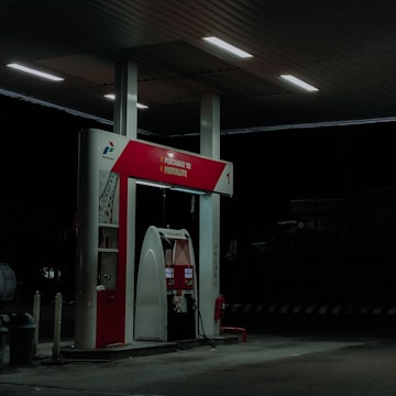 A dimly lit petrol station at night featuring a single fuel pump with the labels Pertamax 92 and Pertalite. The atmosphere is quiet with minimal lighting coming from overhead lights.