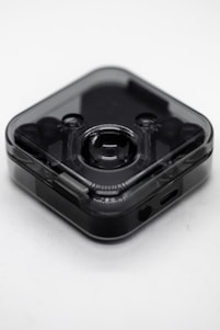 A small, translucent, square-shaped electronic device with rounded edges. It has a central lens-like feature suggesting it could be a camera or a sensor. The device appears to be sleek and modern with a dark, semi-transparent casing. There is a small port at the bottom, likely for charging or data transfer.