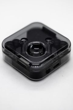 A small, translucent, square-shaped electronic device with rounded edges. It has a central lens-like feature suggesting it could be a camera or a sensor. The device appears to be sleek and modern with a dark, semi-transparent casing. There is a small port at the bottom, likely for charging or data transfer.