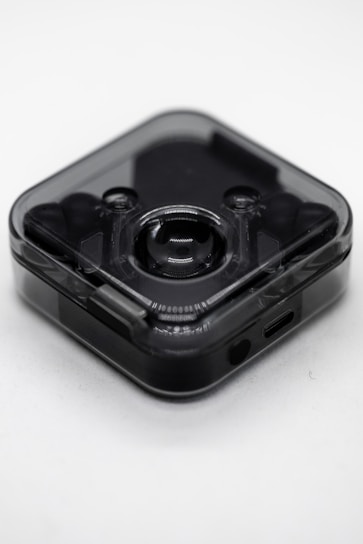 A small, translucent, square-shaped electronic device with rounded edges. It has a central lens-like feature suggesting it could be a camera or a sensor. The device appears to be sleek and modern with a dark, semi-transparent casing. There is a small port at the bottom, likely for charging or data transfer.