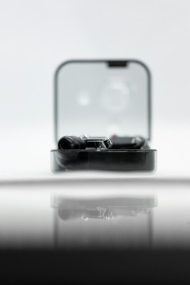 A sleek black gadget is partially enclosed in a clear rectangular case, reflecting on a glossy surface. The backdrop is soft and out-of-focus, creating a minimalist and modern aesthetic.