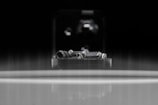 A close-up of an intricate metal mechanism sits within a reflective environment, featuring small, polished components, potentially part of a machine or device. The background is dark, creating a strong contrast against the metallic elements.