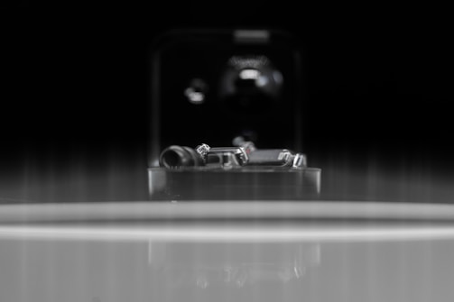 A close-up of an intricate metal mechanism sits within a reflective environment, featuring small, polished components, potentially part of a machine or device. The background is dark, creating a strong contrast against the metallic elements.