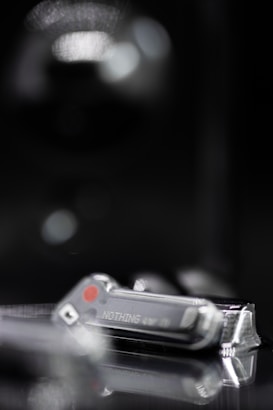 A close-up of a small, transparent object with a metallic appearance, bearing the word 'NOTHING' on its surface. It is placed on a reflective surface, suggesting a high-tech or futuristic theme. The background is blurry and dark, highlighting the object.