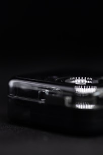 A close-up view of a transparent, rectangular object with rounded edges, set against a dark background. The object reflects light, displaying a series of intricate, concentric patterns. The lighting creates a stark contrast between the object and the shadowy surroundings.