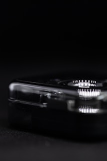A close-up view of a transparent, rectangular object with rounded edges, set against a dark background. The object reflects light, displaying a series of intricate, concentric patterns. The lighting creates a stark contrast between the object and the shadowy surroundings.