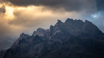 A rugged mountain range under a stormy sky, with a small campfire glowing below.