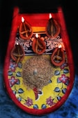 A decorative platter with a colorful floral and patterned design, featuring five traditional oil lamps lit by small flames. The lamps are arranged around a central mound of grains with a small conch shell placed on top. The colors of the platter include bright red, yellow, and blue, with intricate floral details.