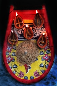 A decorative platter with a colorful floral and patterned design, featuring five traditional oil lamps lit by small flames. The lamps are arranged around a central mound of grains with a small conch shell placed on top. The colors of the platter include bright red, yellow, and blue, with intricate floral details.