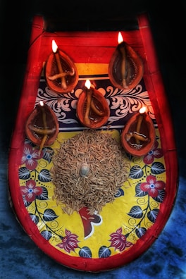 A decorative platter with a colorful floral and patterned design, featuring five traditional oil lamps lit by small flames. The lamps are arranged around a central mound of grains with a small conch shell placed on top. The colors of the platter include bright red, yellow, and blue, with intricate floral details.