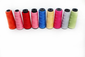 A row of nine spools of thread in various colors are arranged neatly on a plain white background. The colors of the threads include orange, pink, red, blue, yellow, magenta, gray, and green. Each spool is cylindrical with black tops and bottoms.