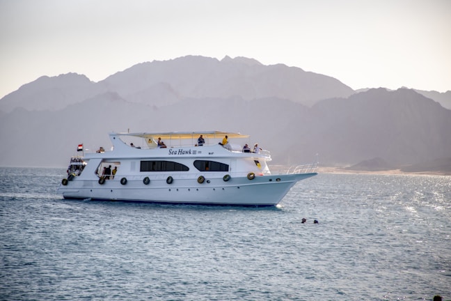 A white yacht named Sea Hawk III is cruising in calm waters with several people visible on its decks. In the background, there are majestic, hazy mountains, and the sun creates a soft glow on the landscape. Two swimmers are in the water near the yacht.