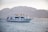 A white yacht named Sea Hawk III is cruising in calm waters with several people visible on its decks. In the background, there are majestic, hazy mountains, and the sun creates a soft glow on the landscape. Two swimmers are in the water near the yacht.