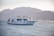 A white yacht named Sea Hawk III is cruising in calm waters with several people visible on its decks. In the background, there are majestic, hazy mountains, and the sun creates a soft glow on the landscape. Two swimmers are in the water near the yacht.