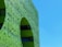 A modern building facade with intricate, abstract green patterns and geometric shapes. The design features a large arch and a combination of smooth and textured surfaces that create a dynamic visual effect under a clear blue sky.